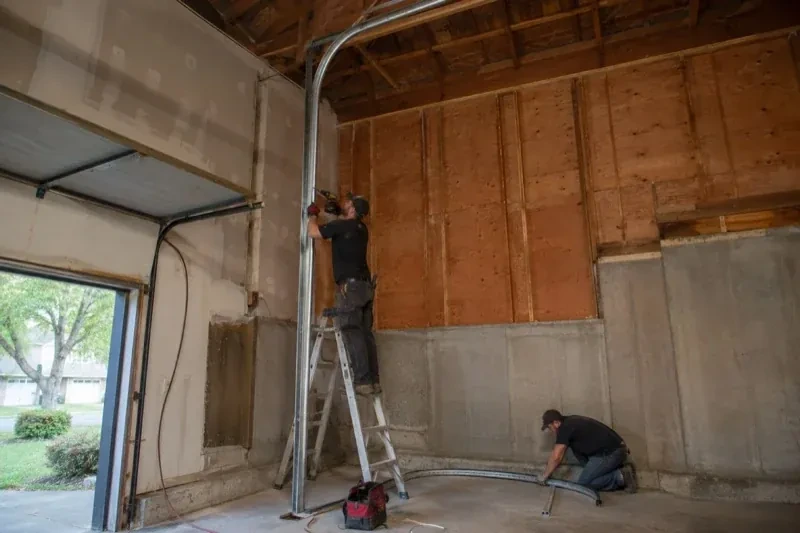 One person on a stepladder working beside a tall curved garage-door track while another kneels beside loose track sections in an unfinished garage. — High-Lift Garage Door Conversion, Woodstock