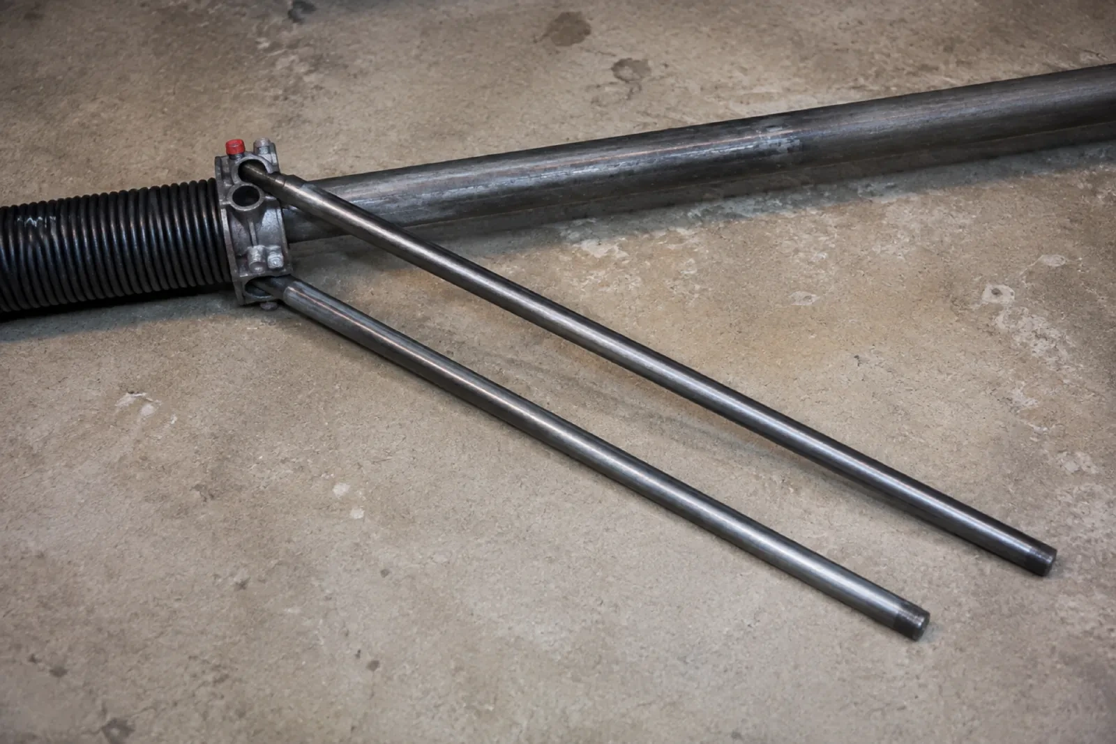 Two long steel winding bars inserted into the cone of a black torsion spring lying on a concrete floor.